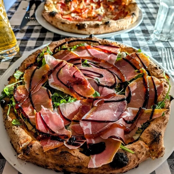 A Charred Brick Oven pizza with prosciutto, arugula, and balsamic glaze, with another pizza in the background.