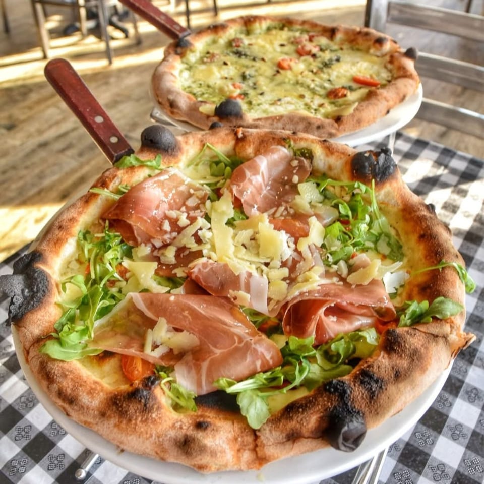 Two Charred Brick Oven pizzas on a checkered tablecloth, one with prosciutto and arugula, the other pesto.