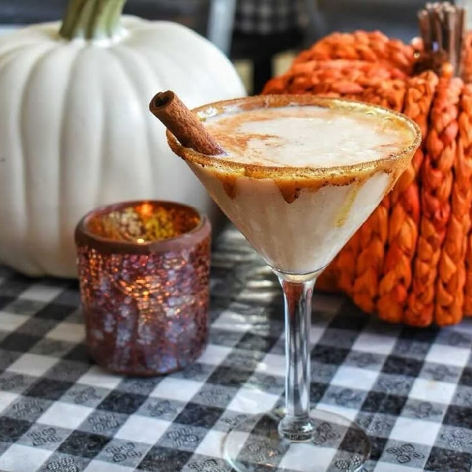 Pumpkin spice martini with a cinnamon stick, next to a lit candle and decorative pumpkins.