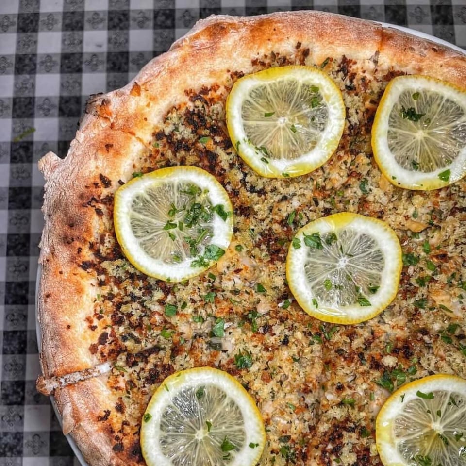 Charred Brick Oven pizza topped with breadcrumbs, parsley, and lemon slices on a checkered tablecloth.
