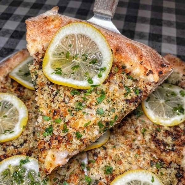 A slice of Charred Brick Oven pizza with a crispy crust, topped with breadcrumbs, parsley, and lemon slices.