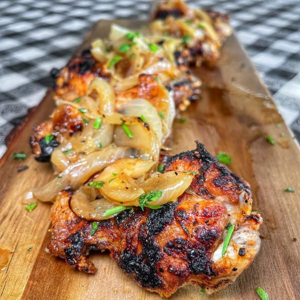 Charred chicken thighs topped with caramelized onions and fresh herbs on a wooden board.