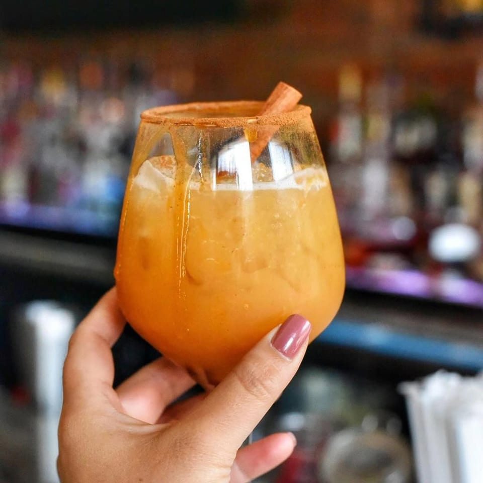 Hand holding an orange cocktail with ice, cinnamon stick, and cinnamon sugar rim at Charred Brick Oven.