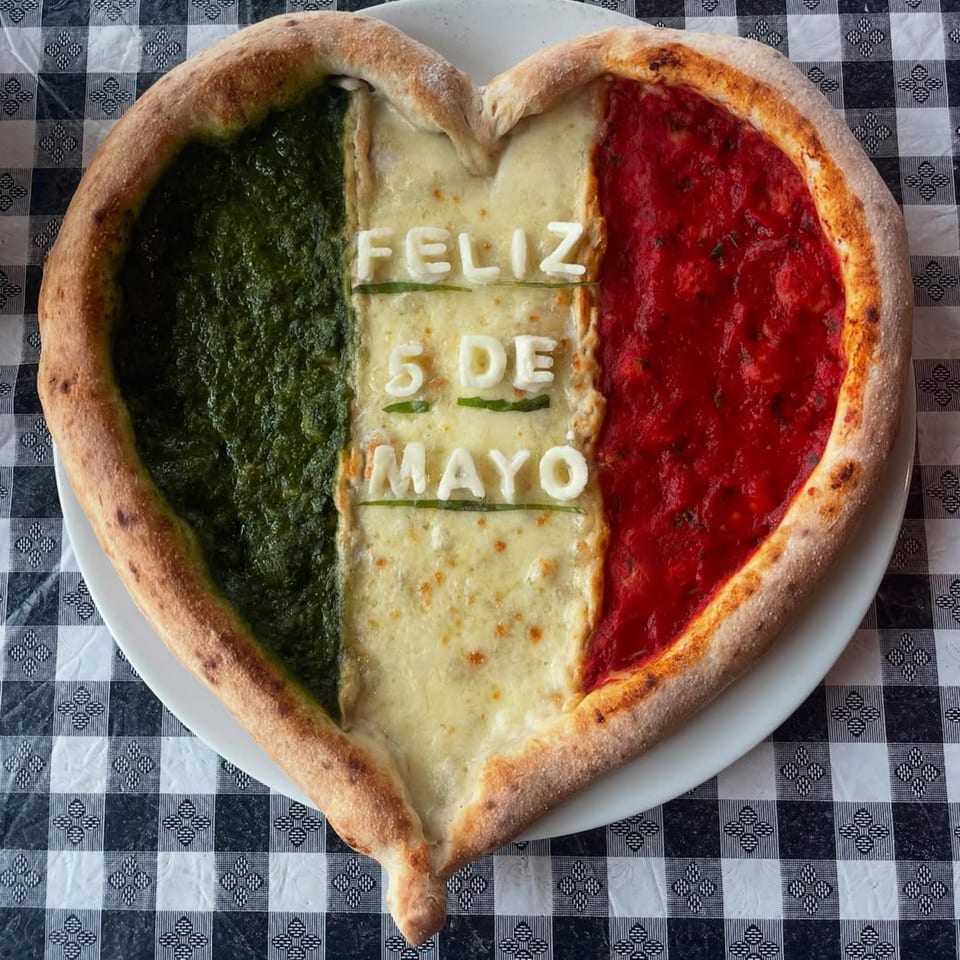 Heart-shaped pizza with green, white, and red toppings, spelling "FELIZ 5 DE MAYO" on a checkered tablecloth.