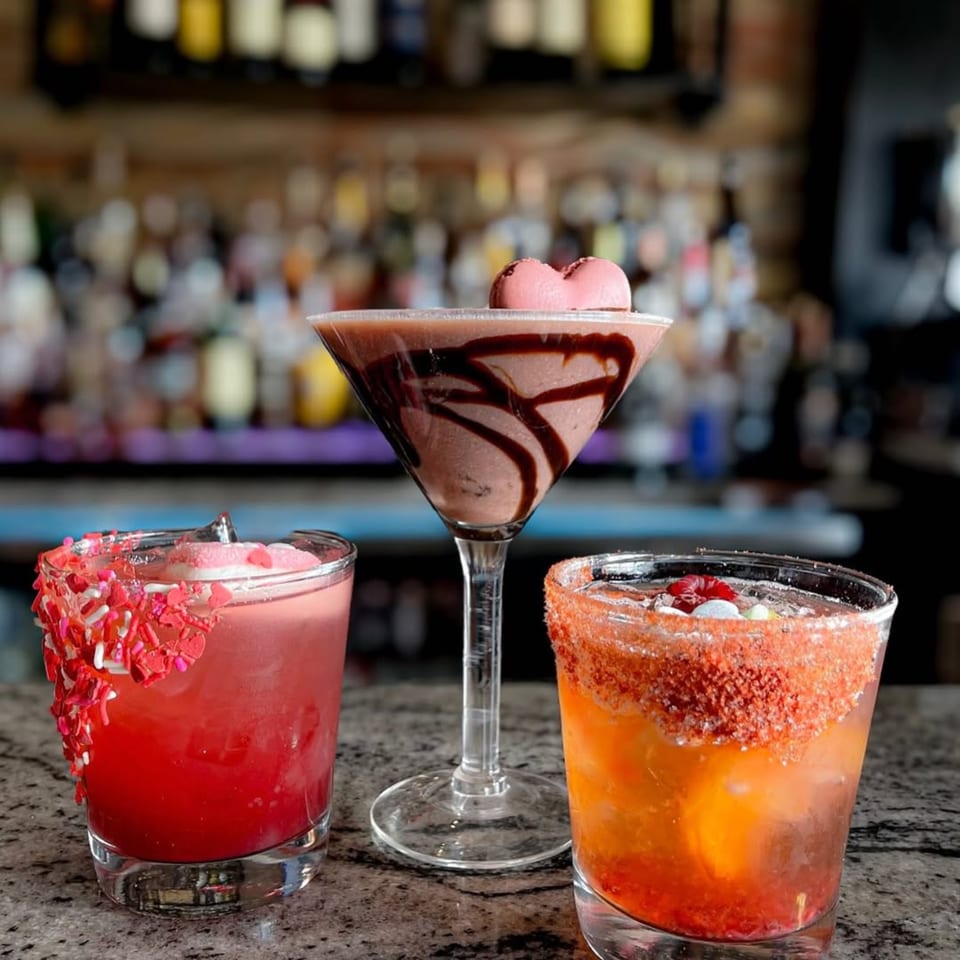 Three colorful cocktails with festive garnishes, including sprinkles and a heart-shaped macaron, on a bar.