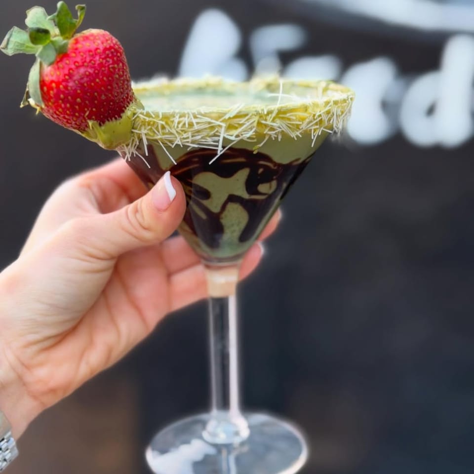 Hand holding a Charred Brick Oven green martini with chocolate drizzle, shredded coconut rim, and strawberry.